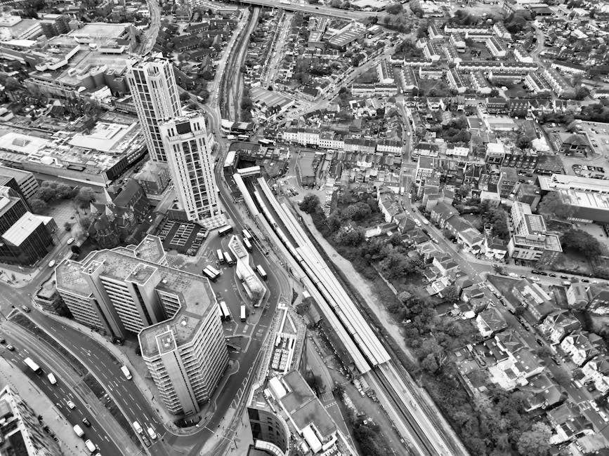 Black and white aerial photograph of a cityscape showing densely packed buildings, including a tall high-rise with a modern design, and several mid-rise office blocks with flat roofs. A railway line w