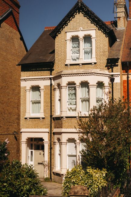 A three-storey Victorian-style house with a beige brick facade, featuring decorative white window frames and a small front garden with bushes and trees. The ground floor has a white entrance door with