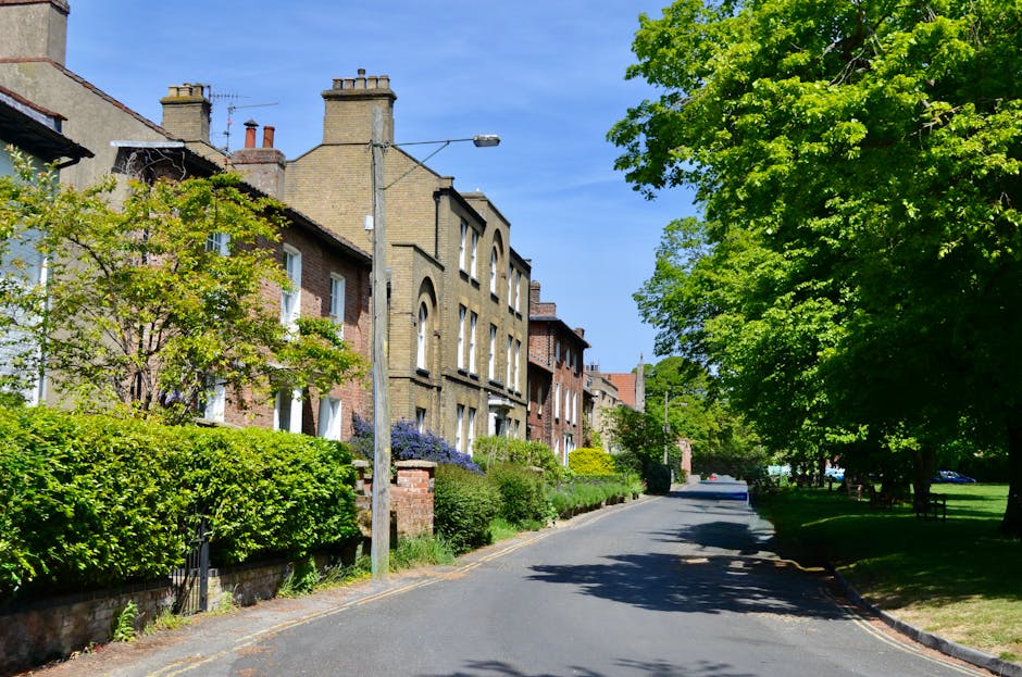 A three-storey Victorian-style house with a beige brick facade, featuring decorative white window frames and a small front garden with bushes and trees. The ground floor has a white entrance door with